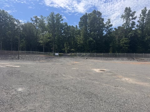 Empty asphalt parking lot or sports field surrounded by chain-link fence with dense green trees and blue sky in background