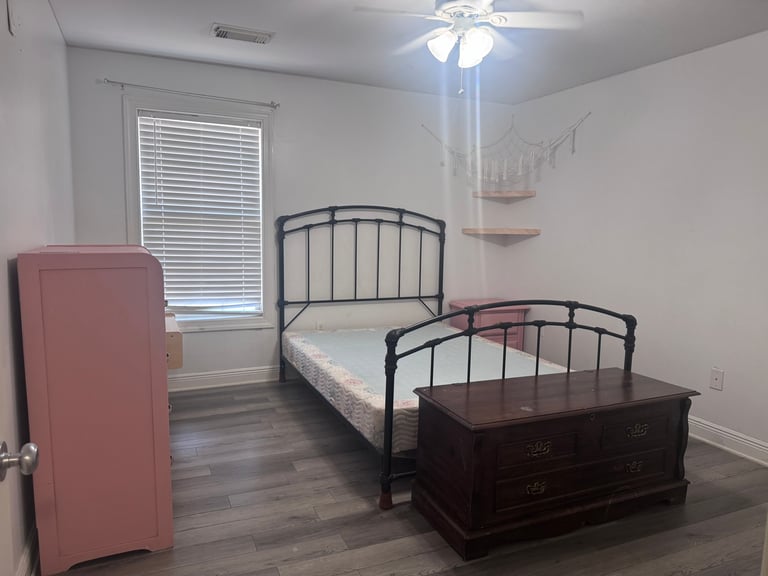Bright bedroom with twin metal frame bed, dark wooden chest, pink cabinet, window with blinds, and ceiling fan.