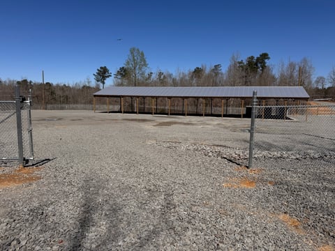 Metal horse stables and paddock enclosure in rural setting with bare ground and trees in background