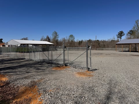Metal gate entrance to a rural gravel farm or ranch area with wooden structures and bare trees under clear blue sky