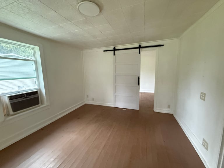 Empty room with wooden flooring, white walls, window on left side, and sliding barn doors on right wall