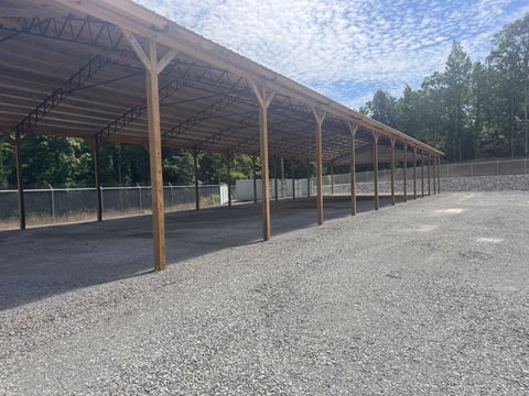 Long wooden pole barn with open sides and metal roof covering a gravel ground area surrounded by trees