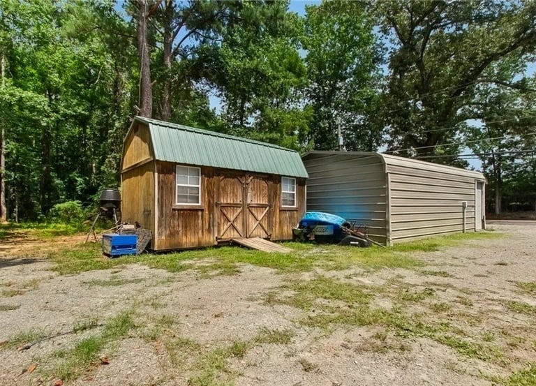 Rural property with wooden barn and metal garage structure surrounded by trees, blue tractor parked between buildings