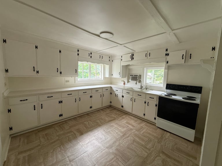 Empty kitchen with white cabinets, stainless steel stove, herringbone tile flooring, and windows above countertop