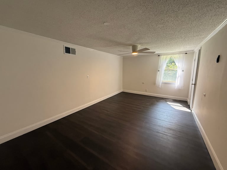 Empty room with dark hardwood floors, white walls, ceiling fan, and window with natural light