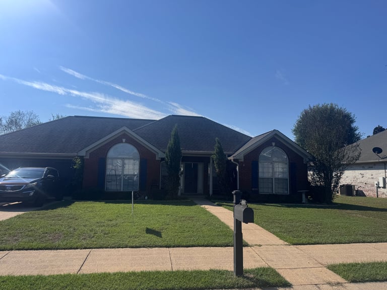 Red brick residential home with dark roof, manicured lawn, driveway, and cypress trees under clear blue sky