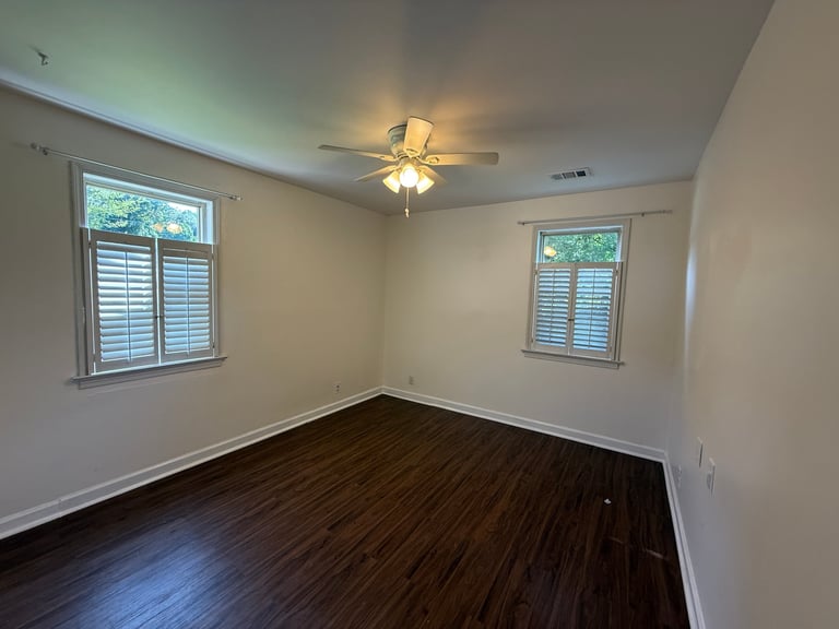 Empty room with white walls, dark wood flooring, ceiling fan, and two windows with shutters