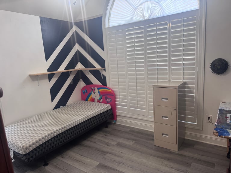 A modern bedroom with a low platform bed featuring a geometric blue and white wall mural headboard, wooden dresser, white shutters, and dark hardwood flooring.