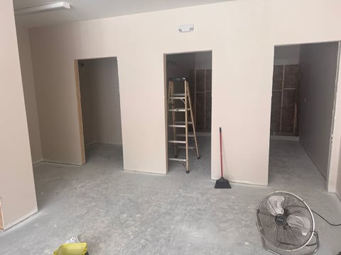Unfinished commercial space with white walls, concrete floor, wooden ladder, floor fan, and construction equipment — new bathroom and shower facilities under construction