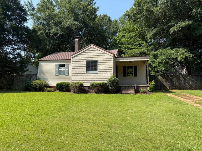 Single-story brick and siding house with front porch and chimney, surrounded by mature trees and a manicured lawn