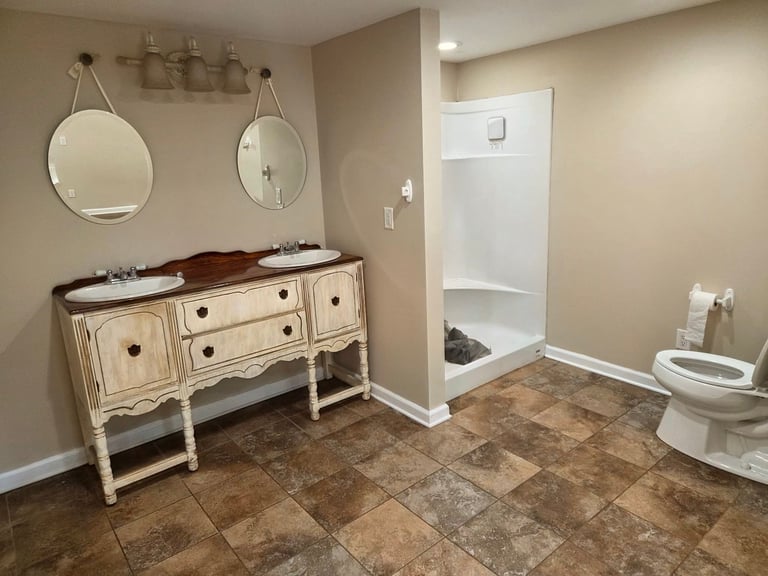 Bathroom with cream-colored vanity, two round mirrors above sink, walk-in shower, toilet, and tile flooring