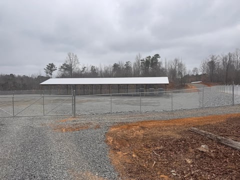 Metal fence enclosing gravel lot with farm buildings and bare trees under cloudy sky