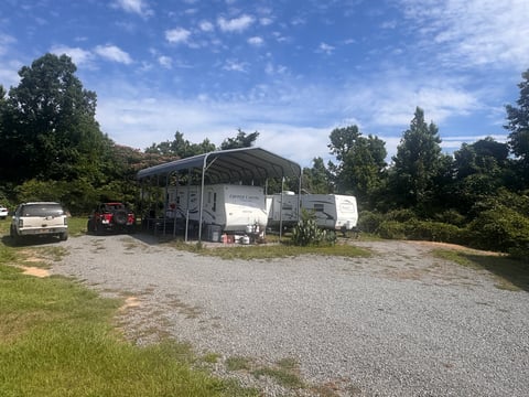 Mobile home with metal carport in rural setting surrounded by trees, with parked vehicles nearby