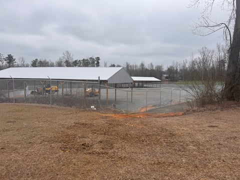 Fenced agricultural area with metal structures and bare ground surrounded by winter trees and overcast sky