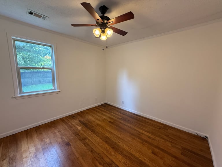 Empty room with wooden floor, white walls, ceiling fan with light fixture, and window with blue exterior view