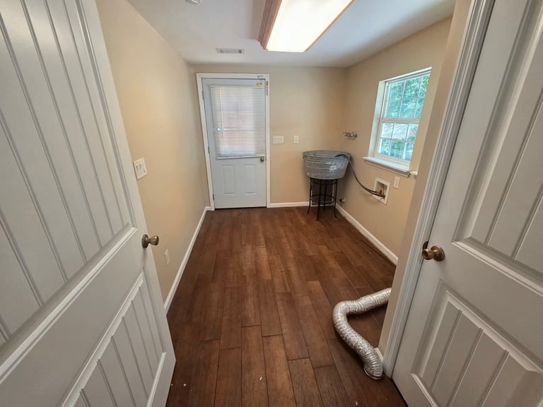 Empty laundry room with wooden flooring, cream walls, white doors, and a window with blinds