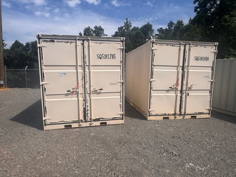 Two beige shipping containers with identification numbers displayed on an outdoor gravel lot with trees in background