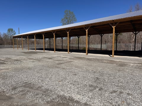 Long wooden pavilion with open-sided roof structure on gravel ground under clear blue sky