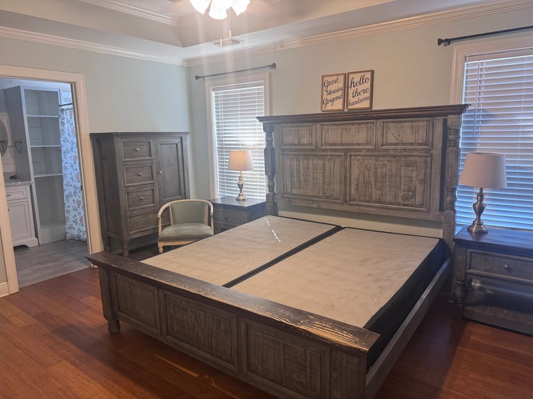 Spacious bedroom with large wooden bed frame, matching dresser and nightstand, hardwood floors, windows with blinds, and ceiling light fixture.