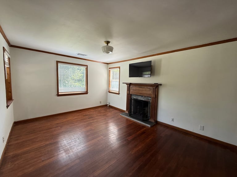 Empty living room with hardwood floors, fireplace, wall-mounted TV, and large windows with wooden trim