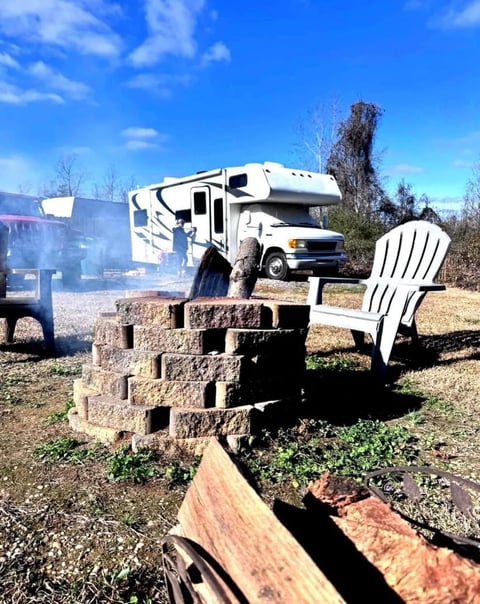 RV campsite with white motorhome, stone fire pit, white adirondack chair, and clear blue sky