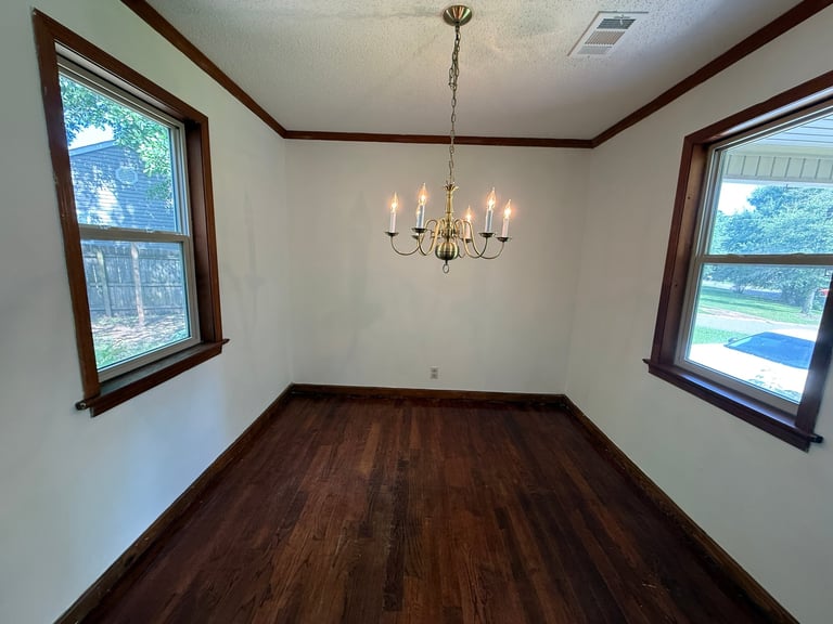Empty room with dark hardwood floors, beige walls, brass chandelier, and two large windows with dark wood frames overlooking green landscape