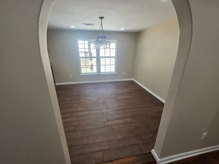 Empty room with dark wood flooring, white walls, and large window with chandelier overhead