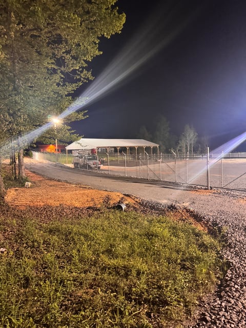 Nighttime outdoor facility with metal-roofed structure and fencing, illuminated by bright floodlights, surrounded by trees and gravel