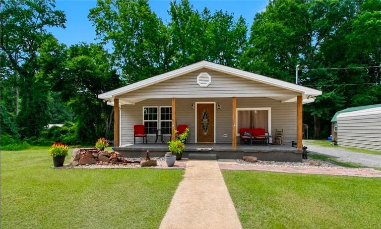 Single-story beige house with front porch, landscaping, and concrete walkway surrounded by green lawn and trees
