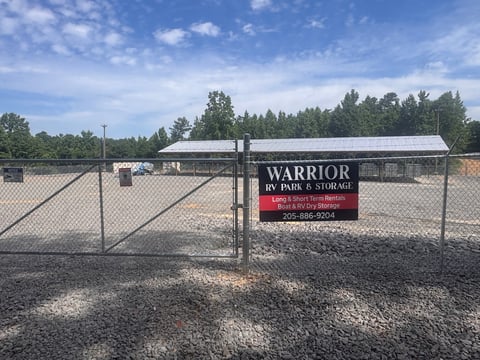 Chain-link fenced storage facility with Warrior RV Park & Storage sign and metal buildings under blue sky