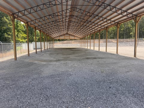Large open-air metal structure with truss roof and gravel floor, surrounded by wire fencing and trees