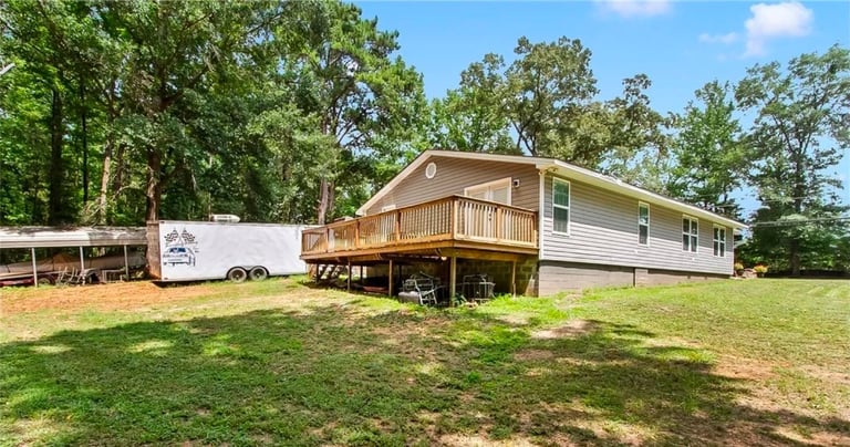 Single-wide mobile home elevated on concrete blocks in a rural setting with green grass and trees, accompanied by a white storage trailer