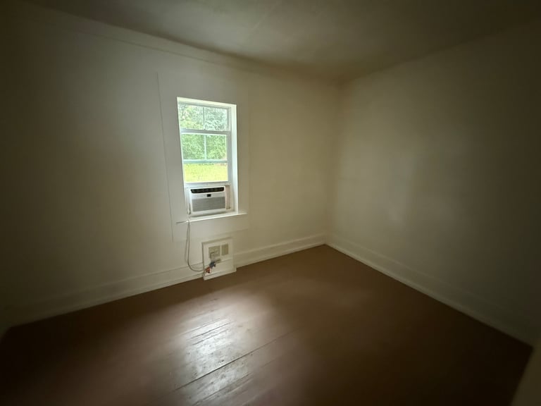 Empty room with cream-colored walls, hardwood floor, window with air conditioning unit, and electrical outlet