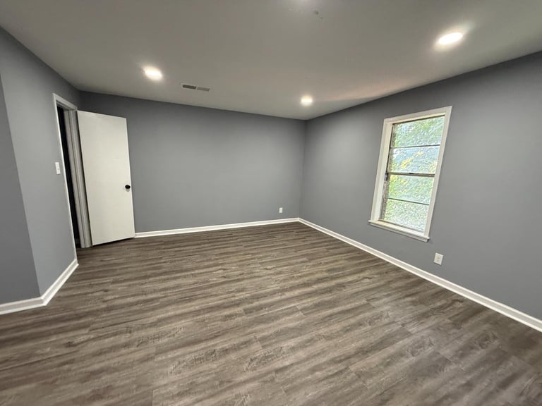 Empty gray room with wood-look flooring, recessed ceiling lights, a white door on the left, and a window on the right wall
