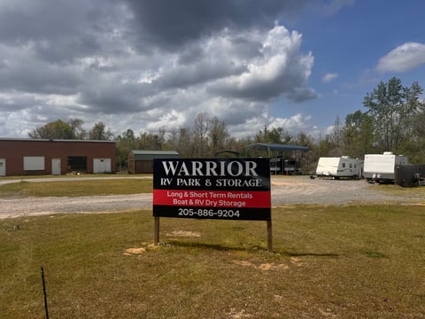 Warrior RV Park and Storage sign in a grassy field with brick building and storage trailers visible in the background under a cloudy sky