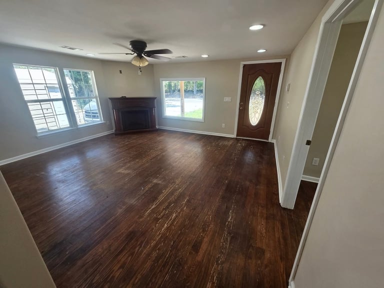 Empty living room with dark wood flooring, ceiling fan, windows, and doorway with decorative oval window