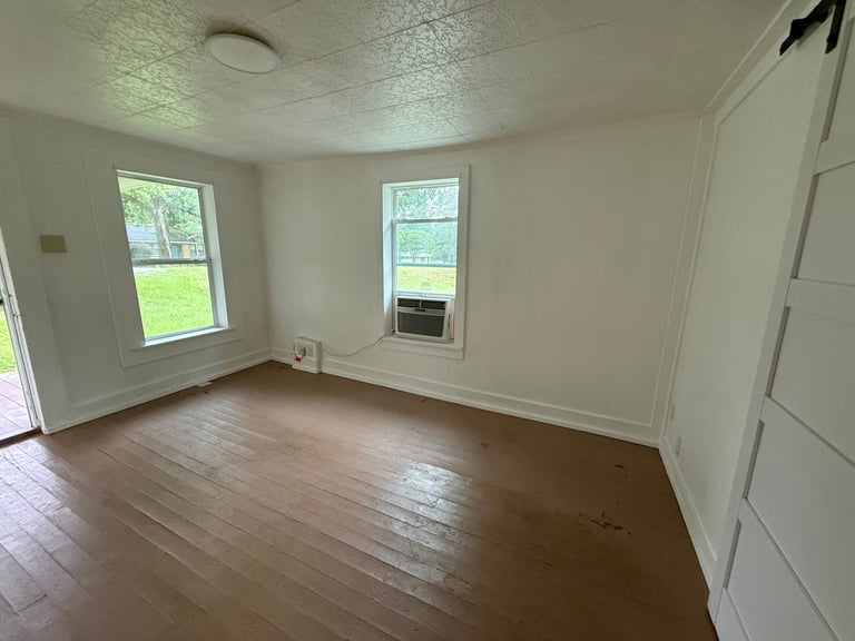 Empty room with hardwood floors, white walls, two windows, and a white door visible on the right side