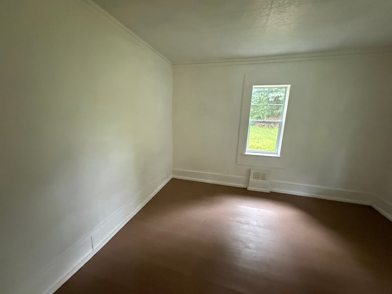 Empty room with white walls, dark hardwood floor, and a window with natural light streaming in