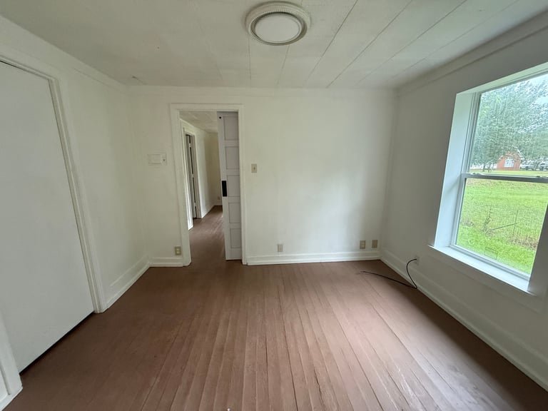 Empty room with dark wood flooring, white walls, recessed ceiling light, doorway to hallway, and large window with outdoor view