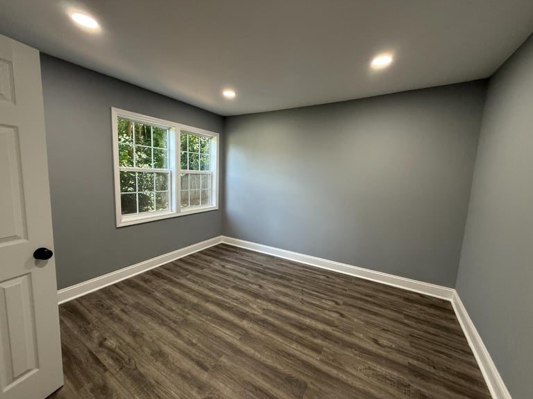 Empty room with gray walls, wood plank flooring, white trim, recessed ceiling lights, and a window overlooking trees