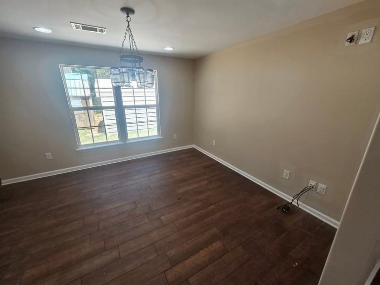 Empty room with hardwood floors, beige walls, large window with shutters, and pendant light fixture hanging from ceiling