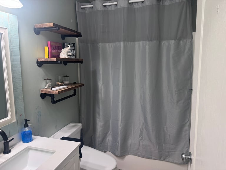 Modern bathroom with gray shower curtain, white sink, toilet, and three floating wooden shelves mounted on the wall