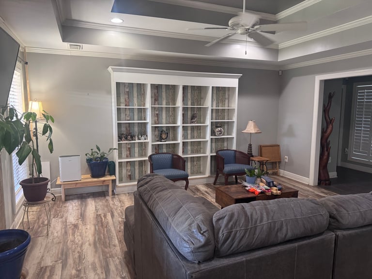 Modern living room with gray sectional sofa, white glass-front bookcase, armchairs, ceiling fan, wood-look flooring, and potted plants