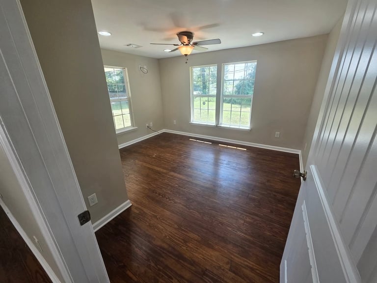 Empty room with dark hardwood floors, white walls, ceiling fan, and large windows overlooking green lawn