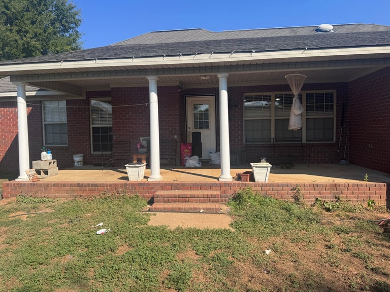 Red brick house with covered porch, white columns, and overgrown front yard under clear blue sky