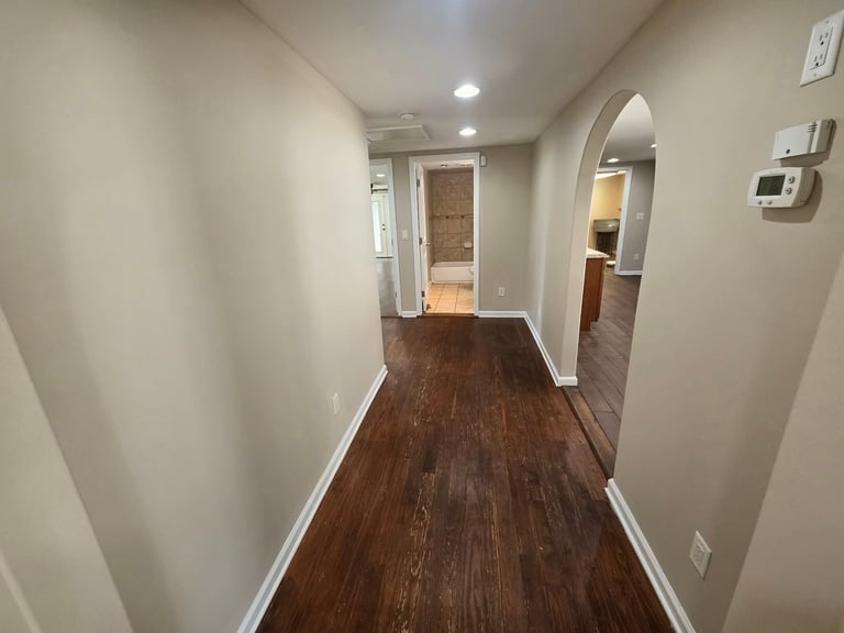Empty hallway with beige walls, dark wood flooring, recessed ceiling lights, and doorways leading to other rooms