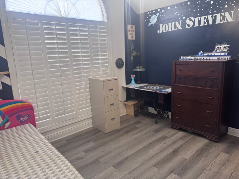 A bedroom interior with white shutters, dark blue accent wall with JOHN STEVEN text, wooden dresser, and nautical decor elements