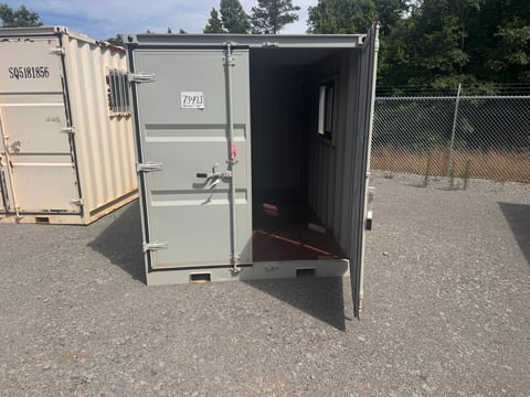Open shipping container with dark interior door on gravel ground, chain-link fence and trees visible in background
