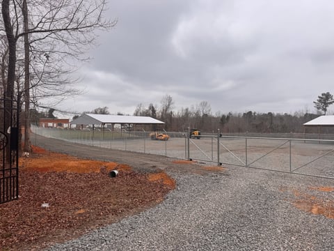 Rural farm property with metal fencing, gravel driveway, scattered buildings, and bare winter trees under cloudy sky